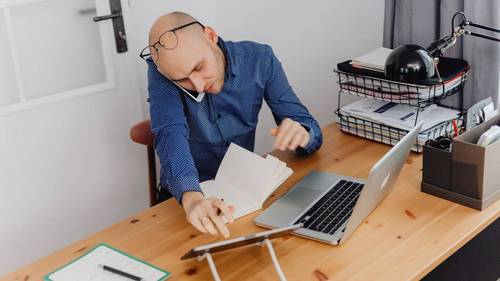 Selbstständiger telefoniert am Schreibtisch und blättert dabei in Unterlagen, daneben Laptop und Ablagekörbe mit Papieren.