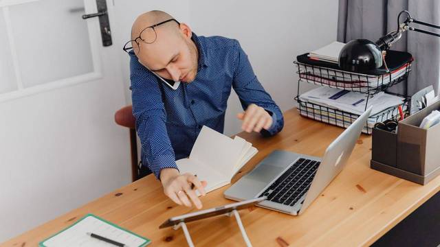 Selbstständiger telefoniert am Schreibtisch und blättert dabei in Unterlagen, daneben Laptop und Ablagekörbe mit Papieren.