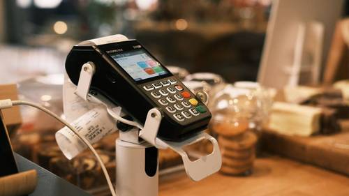 Kartenzahlungsterminal auf einem weißen Standfuß am Tresen eines Cafés, daneben eine Bonrolle mit ausgedruckten Belegen, im unscharfen Hintergrund Gebäck und Auslage.