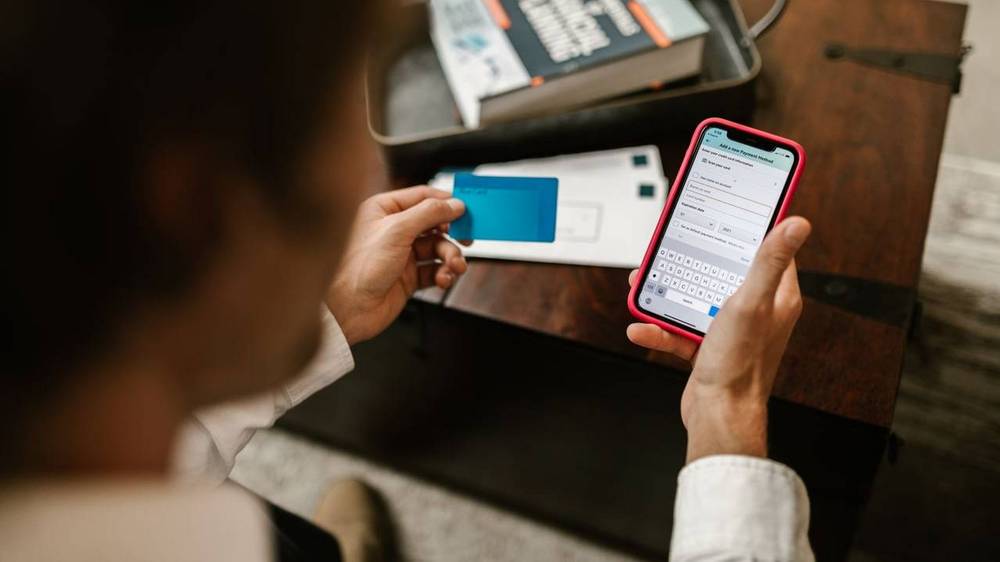 Person hält ein Smartphone mit einem Eingabeformular für eine neue Zahlungsmethode und in der anderen Hand eine blaue Bankkarte, im Hintergrund ein Buch zur Finanzplanung.