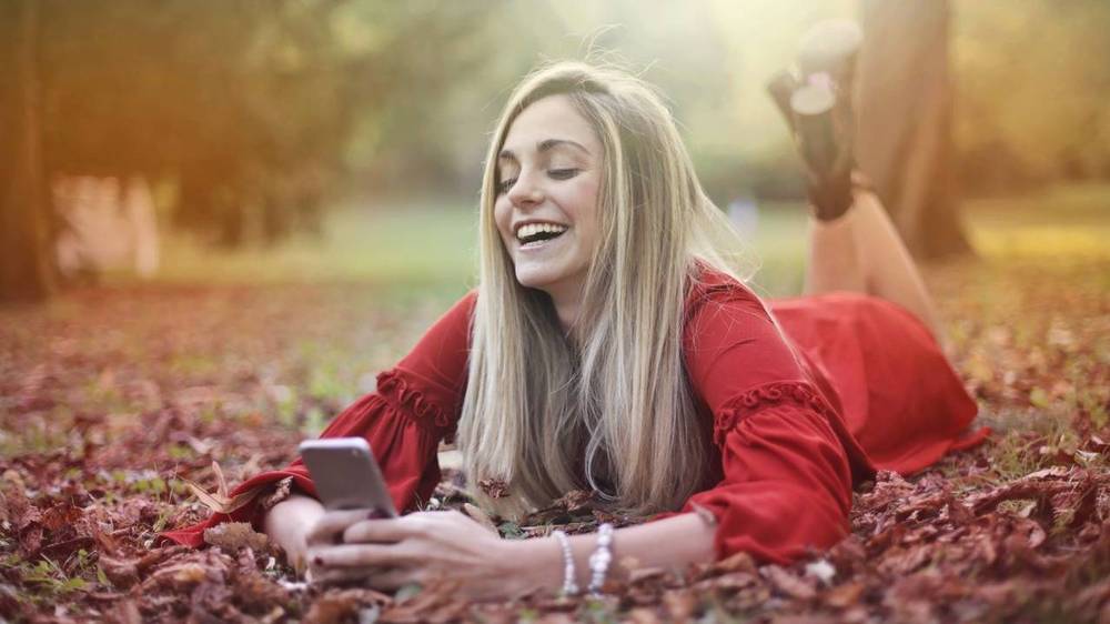 Lachende Frau im roten Kleid liegt im herbstlichen Laub und schaut auf ihr Smartphone.