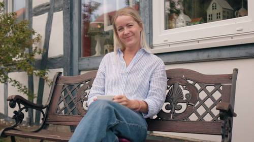 Marie-Theres Mund sitzt mit einer Kaffeetasse auf einer Holzbank vor einem Fachwerkhaus.