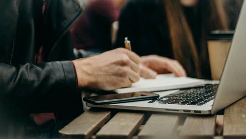 Person sitzt vor Laptop mit Notizblock und Bleistift