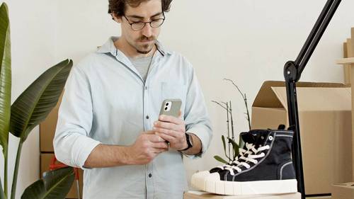 Mann mit Brille schaut in einem kleinen Lagerraum zwischen Kartons auf sein Smartphone.