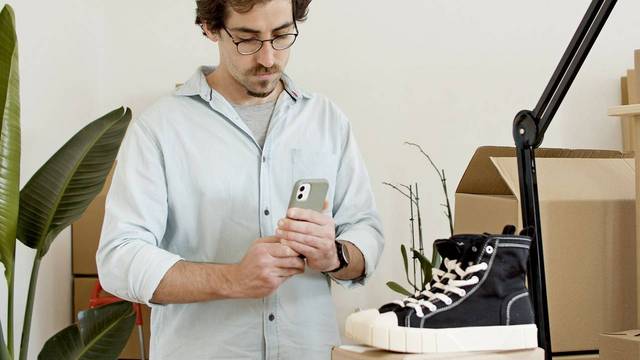 Mann mit Brille schaut in einem kleinen Lagerraum zwischen Kartons auf sein Smartphone.