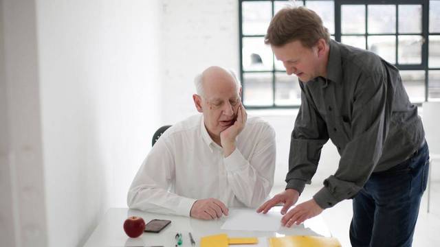 Unternehmensnachfolge Ein älterer Mann sitzt an einem Schreibtisch, ein jüngerer Mann steht daneben; beide sehen auf Unterlagen und Aktenmappen.