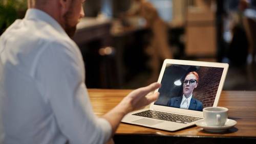 Mann von hinten an einem Laptop in einem Café, auf dem Bildschirm eine Frau im Business-Outfit während eines Video-Calls.